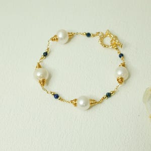May include: A gold bracelet with large, white pearls and small, dark blue beads. The pearls are wire-wrapped in gold, and the bracelet has a decorative clasp. The jewellery is set against a light background.
