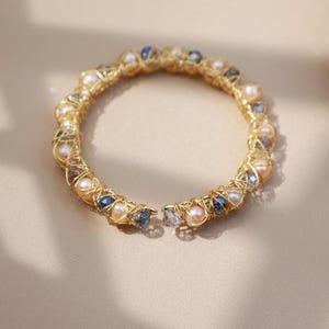 May include: A gold bracelet with a woven design, featuring pearls and blue and peach-coloured beads. The bracelet is displayed on a neutral surface. The bracelet's design is intricate.