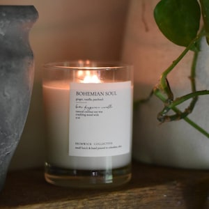 May include: A clear glass candle jar with a white label that reads "BOHEMIAN SOUL" and "bae fragrance candle". The candle is lit and the wax is melting. The label also includes the scent notes "ginger, vanilla, patchouli" and the brand name "BRIMWICK COLLECTIVE".