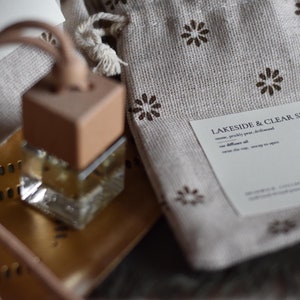 May include: A small, clear glass bottle with a wooden cap and a brown leather cord. The bottle is filled with a light yellow liquid. The bottle is sitting on a gold tray. The bottle is labeled "Lakeside & Clear Skies" with the text "notes: prickly pear, driftwood, our diffuser oil, warm the cap, snap in open".
