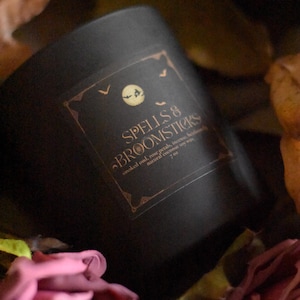 May include: A black candle with a gold label that reads "SPELLS & BROOMSTICKS." The label includes the ingredients: smoked oud, rose petals, and incense. The candle is 7 oz. and made with natural coconut soy wax. The image includes pink roses.