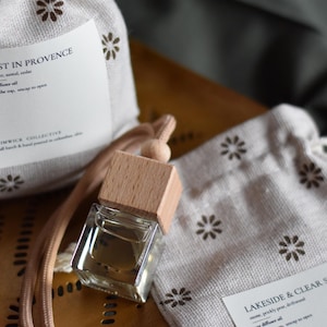 May include: Two small white fabric pouches with brown floral patterns and labels that read "Lost in Provence" and "Lakeside & Clear Skies". A clear glass bottle with a wooden cap is attached to a brown cord.