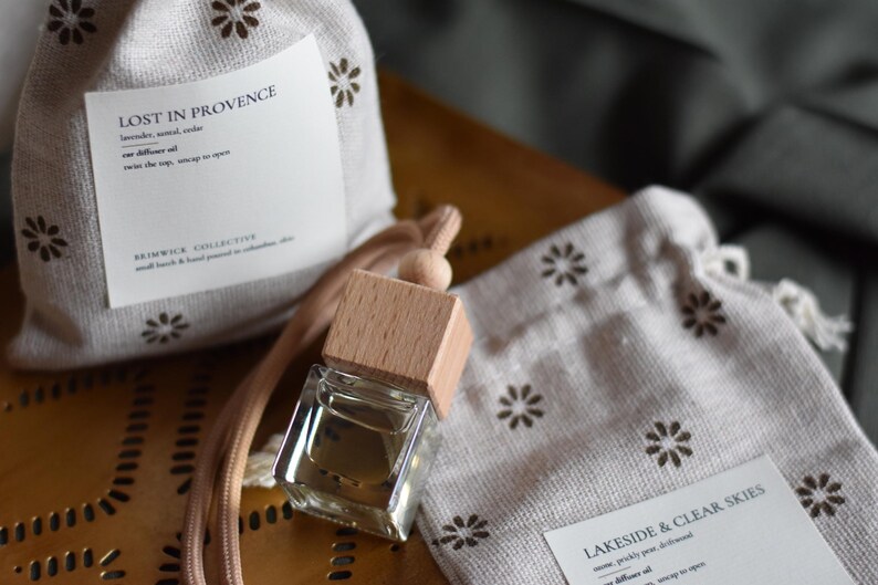 May include: Two small white fabric pouches with brown floral patterns and labels that read "Lost in Provence" and "Lakeside & Clear Skies". A clear glass bottle with a wooden cap is attached to a brown cord.