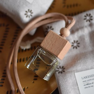May include: A wooden car air freshener with a clear glass bottle and a brown cord. The air freshener is on a brown surface with a white fabric with a floral pattern.