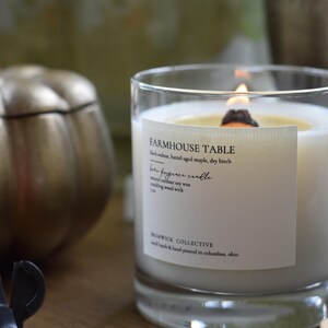 May include: A lit candle in a clear glass jar with a label that reads "FARMHOUSE TABLE". The candle has a wooden wick and is placed on a wooden surface. A gold pumpkin-shaped container is in the background.