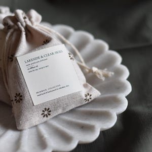 May include: Two small white fabric pouches with brown floral designs. The pouches are tied with string and contain small labels with the text "Lakeside & Clear Skies" and "Brunswick Collective, small batch & hand poured in collaboration, etc."