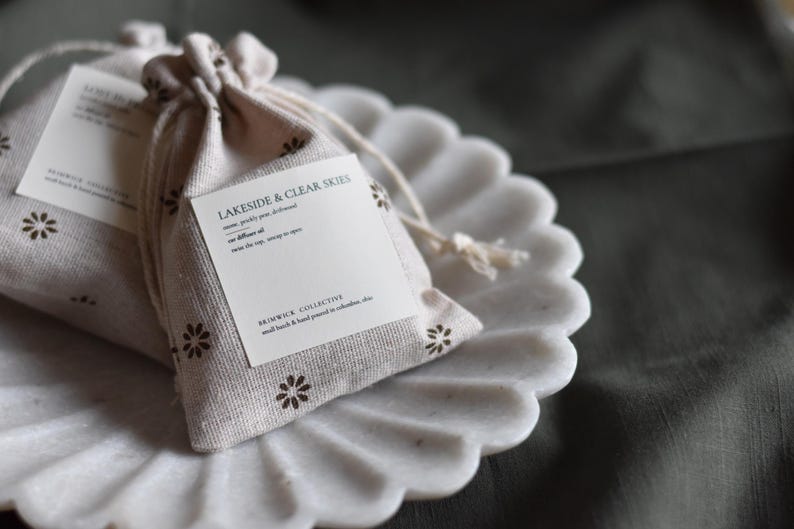 May include: Two small white fabric pouches with brown floral designs. The pouches are tied with string and contain small labels with the text "Lakeside & Clear Skies" and "Brunswick Collective, small batch & hand poured in collaboration, etc."