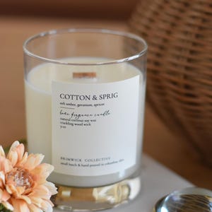 May include: A white candle in a clear glass jar with a gold rim. The label reads "Cotton & Sprig" and "Home Fragrance Candle". The candle is a natural coconut soy wax with a crackling wood wick. The candle is 9 oz.