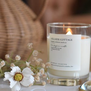 May include: A white candle in a glass jar with a label that reads "Hillside Cottage" and "Home Fragrance Candle". The candle is lit and the flame is visible. The candle is sitting on a book with the word "LIFESTYLED" printed on the cover.