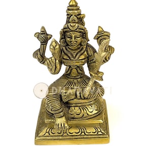 May include: A gold-colored brass statue of a Hindu goddess sitting on a square base. The goddess is holding a lotus flower in one hand and a sword in the other. She is wearing a crown and elaborate jewelry.