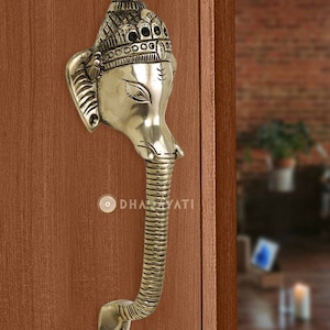 May include: Brass door handle featuring a detailed Ganesha head with a textured trunk and a decorative crown. The handle is mounted on a wooden door.