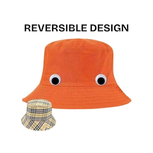 Peut inclure: Chapeau seau orange avec des yeux globuleux et le texte "REVERSIBLE DESIGN". Un chapeau plus petit et réversible avec un motif à carreaux est à côté du chapeau orange. Les chapeaux sont un accessoire amusant et ludique.