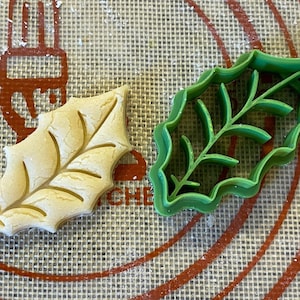 May include: A green plastic holly leaf cookie cutter and a light brown cookie in the shape of a holly leaf on a brown and white silicone baking mat.