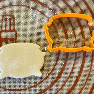 May include: A close-up of a cookie cutter and a cookie dough cutout on a brown and white baking mat. The cookie cutter is orange and shaped like a speech bubble. The cookie dough cutout is a light brown colour and is also shaped like a speech bubble.