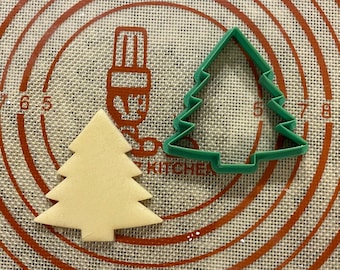 以下が含まれることがあります： 緑色のプラスチック製のクリスマスツリーのクッキー型と、茶色のクリスマスツリーの形をしたクッキーが、茶色と白の模様のベーキングマットの上に置かれています。
