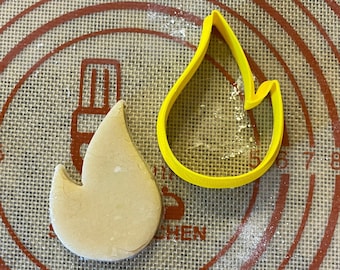 May include: A yellow plastic cookie cutter shaped like a flame, and a light brown cookie in the shape of a flame on a brown and red patterned surface.