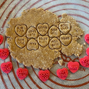 Puede incluir: Primer plano de una estera de hornear de silicona roja con una hoja de masa para galletas marrón encima. La masa para galletas está presionada con cortadores de galletas en forma de corazón con varios mensajes sobre perros, como "Fur Ever", "Good Dog", "Belly Rubs", "Hot Dog", "Roll Over", "Paw", "Top Dog", "Baby Fur", "You Ruff" y "XOXO".