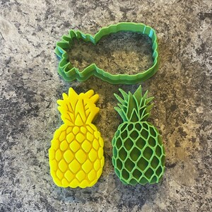 Puede incluir: Tres cortadores de galletas de piña, uno verde y dos amarillos. El cortador verde es un contorno detallado de una piña, mientras que los cortadores amarillos son piñas sólidas con superficies texturizadas.