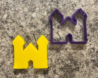 May include: Two cookie cutters in the shape of a castle. The larger cutter is purple and the smaller cutter is yellow.