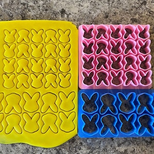 May include: Three bunny rabbit shaped cookie cutters. The cutters are yellow, pink and blue. The yellow cutter is filled with yellow dough, the pink cutter is filled with pink dough and the blue cutter is empty.