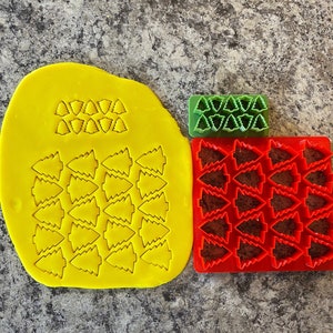 May include: Three yellow silicone cookie cutters shaped like Christmas trees. The cutters are arranged in a row, with the largest cutter in the middle and the two smaller cutters on either side. The cutters are on a grey work surface.