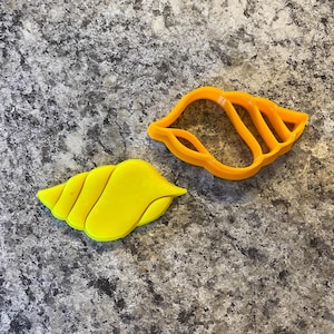 Puede incluir: Dos elementos en forma de concha. Uno es una concha sólida de color amarillo brillante. El otro es un cortador de galletas en forma de concha de color naranja. Ambos están sobre una superficie gris moteada. Los artículos son probablemente para hornear o hacer manualidades.