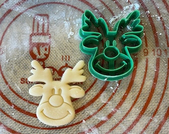 May include: A green plastic reindeer cookie cutter and a beige cookie in the shape of a reindeer on a brown and green baking mat.