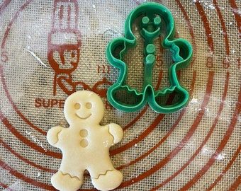 May include: A green gingerbread man cookie cutter and a plain gingerbread man cookie on a brown and white patterned surface.