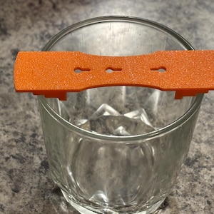 May include: An orange plastic bar with three small holes and two side supports, resting on a clear glass. The bar has a textured surface and is designed to fit over the glass.