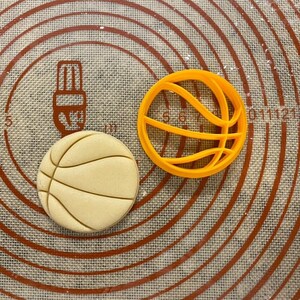 Può includere: Primo piano di due stampini per biscotti a forma di pallacanestro su un tappetino da forno in silicone marrone. Uno stampino è arancione e l'altro è un biscotto marrone chiaro.