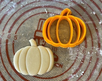 May include: A close-up of a pumpkin-shaped cookie cutter and a pumpkin-shaped cookie. The cookie cutter is orange and the cookie is light brown. Both are on a brown and white patterned surface.