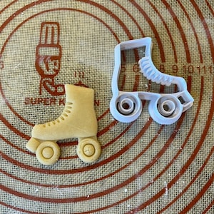 Peut inclure: Un emporte-pièce en plastique blanc en forme de patins à roulettes et un biscuit brun au sucre en forme de patins à roulettes. L'emporte-pièce est sur un tapis de cuisson en silicone blanc avec des cercles rouges.