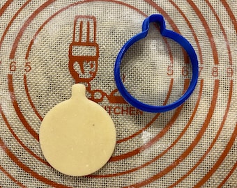 May include: A blue plastic Christmas ornament cookie cutter and a plain round cookie on a red and white silicone baking mat.