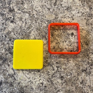 Puede incluir: Un cortador de galletas cuadrado amarillo y un cortador de galletas cuadrado rojo sobre una encimera gris.