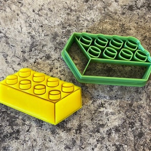 Puede incluir: Dos cortadores de galletas de plástico con forma de bloques de Lego. El cortador más grande es verde y el más pequeño es amarillo.