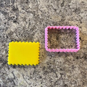 Puede incluir: Dos cortadores de galletas, uno amarillo y otro rosa, ambos con un borde ondulado y una forma rectangular.