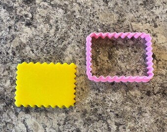 Puede incluir: Dos cortadores de galletas, uno amarillo y otro rosa, ambos con un borde ondulado y una forma rectangular.
