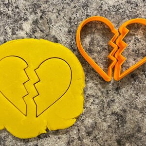 May include: A close-up of a broken heart cookie cutter. The cutter is orange and is made of plastic. The cutter is on a gray countertop. The cutter is next to a yellow piece of cookie dough that has been cut out with the cutter.