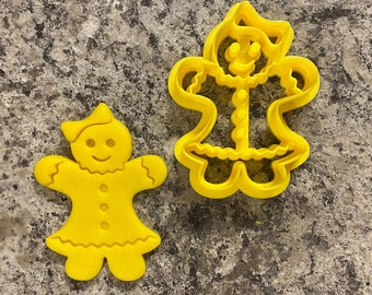 May include: Two yellow gingerbread cookie cutters, one is a traditional gingerbread person shape and the other is a gingerbread person wearing a dress and a hat.