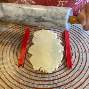 Puede incluir: Un rodillo de mármol blanco extendiendo un círculo de masa sobre una estera de hornear de silicona roja y blanca con guías de medición rojas marcadas con 1/4.