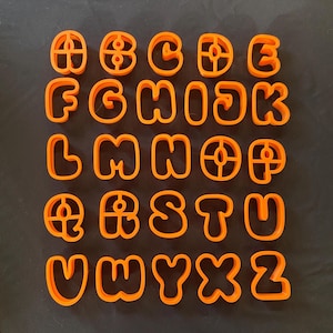 Puede incluir: Cortadores de galletas de plástico naranja con el alfabeto inglés completo de la A a la Z. Cada letra tiene una fuente redondeada y con burbujas. Los cortadores están dispuestos sobre una superficie oscura.