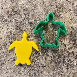 May include: A green plastic turtle cookie cutter and a yellow plastic turtle cookie. The cookie cutter is on the right and the cookie is on the left.