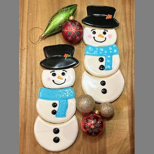 May include: Two decorated gingerbread snowman cookies, one with a blue scarf and the other with a red and white scarf. The cookies are on a wooden surface with red and gold baubles.