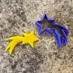 Peut inclure: Deux emportes-pièces, l'un jaune et l'autre violet, en forme d'étoile filante. L'étoile jaune a une forme d'étoile à cinq branches et l'étoile violette a une forme d'étoile à six branches.
