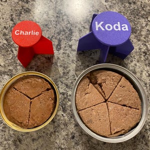 May include: Two cans of wet cat food, one with a red label that says "Charlie" and the other with a blue label that says "Koda".
