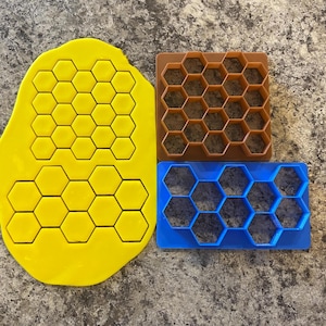 Op de afbeelding: Drie honingraatkoekjesuitstekers, een gele, een bruine en een blauwe. De uitstekers zijn gemaakt van plastic en hebben een zeshoekig ontwerp.