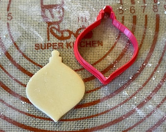 以下が含まれることがあります： 茶色と白のシリコン製のベーキングマットの上に、赤いプラスチック製のクリスマスオーナメントのクッキーカッターが置かれています。オーナメントの形をした、焼かれていないプレーンなクッキーも置かれています。
