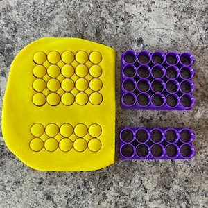 May include: A yellow fondant sheet with two purple plastic cookie cutters. The cookie cutters are shaped like a square and a rectangle. Each cutter has multiple circular cutouts.