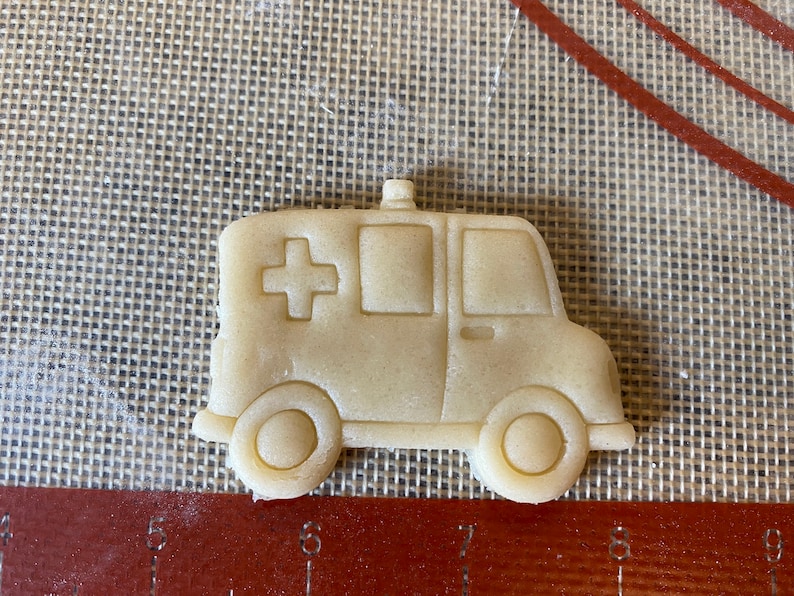 Pu&ograve; includere: Un biscotto di colore marrone chiaro a forma di ambulanza con una croce rossa sul lato. Il biscotto &egrave; su un tappetino da forno marrone.
