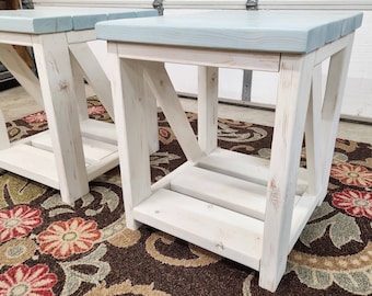 May include: Two white wooden side tables with a light blue top and a lower shelf. The tables have an X-shaped support on the sides. The tables are on a floral patterned rug.